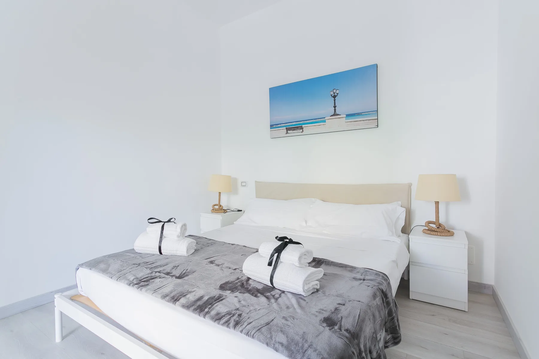 A bright bedroom with a bed, bedside tables, and beach artwork above the headboard.