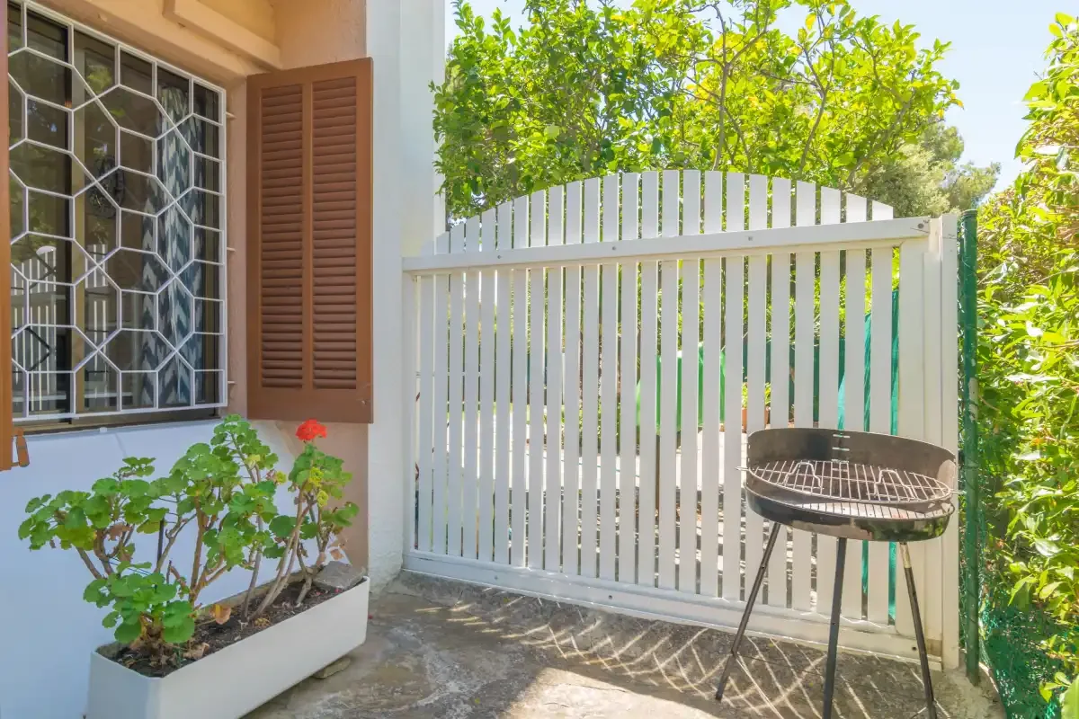 A small outdoor patio has a potted plant beside a white gate and grill.