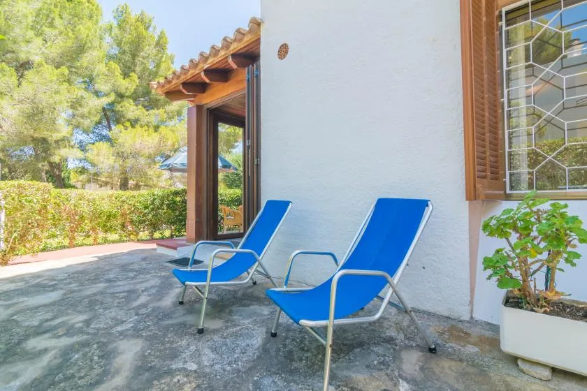 An outdoor patio beside a white wall, with blue seating and leafy trees nearby.