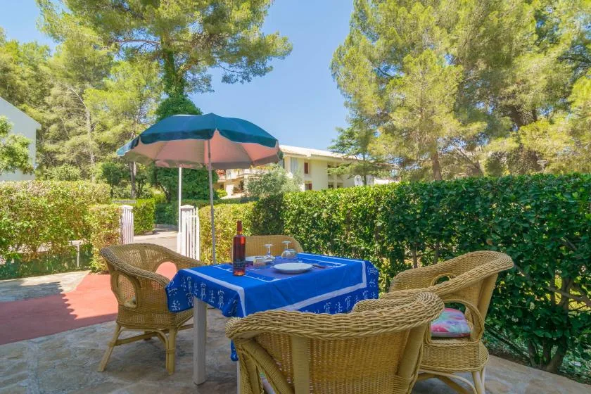 An outdoor dining area with a table under a colorful umbrella, surrounded by greenery.