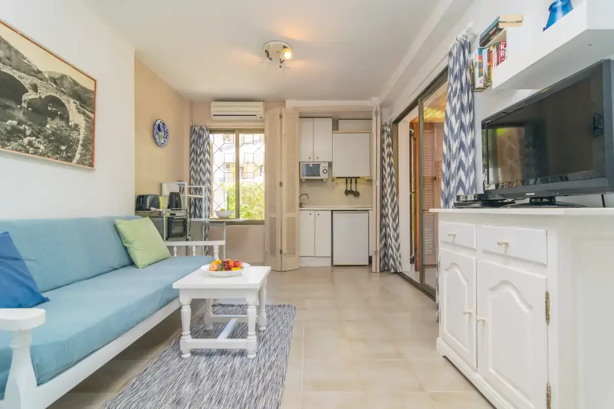 A living room with blue seating and a small white table, opening onto a compact kitchen.