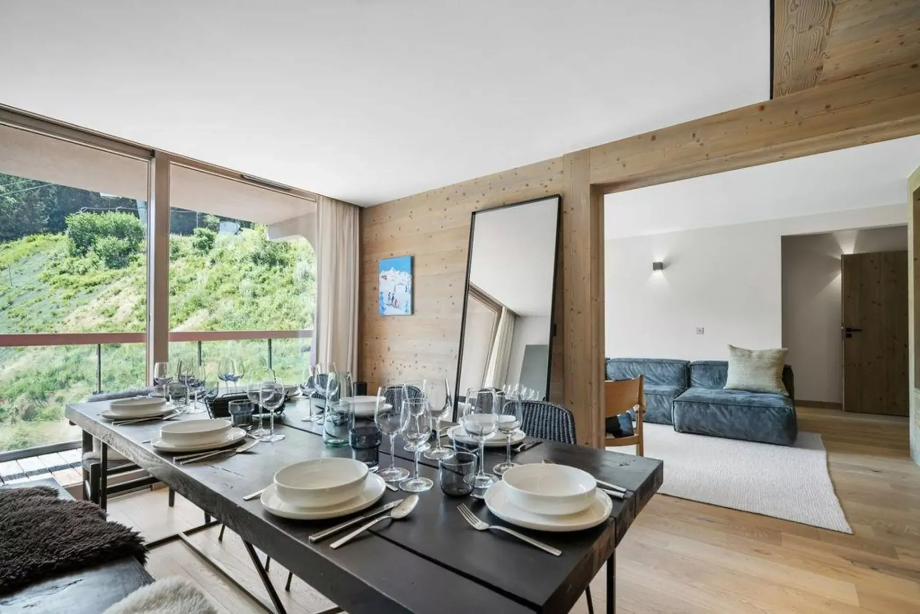 A dining area with a set table beside large windows overlooking greenery.
