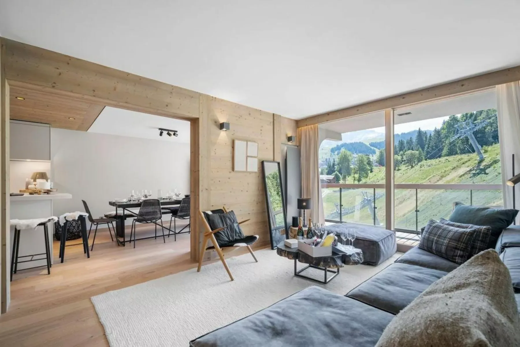 A bright living area opens onto a dining space, with large windows overlooking green hills.
