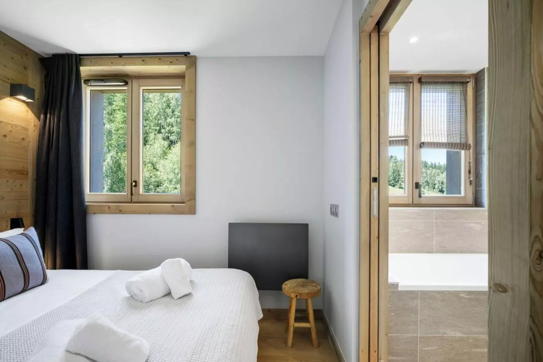 A bedroom with a white bed and a wooden-framed window, with a bathroom visible through the doorway.