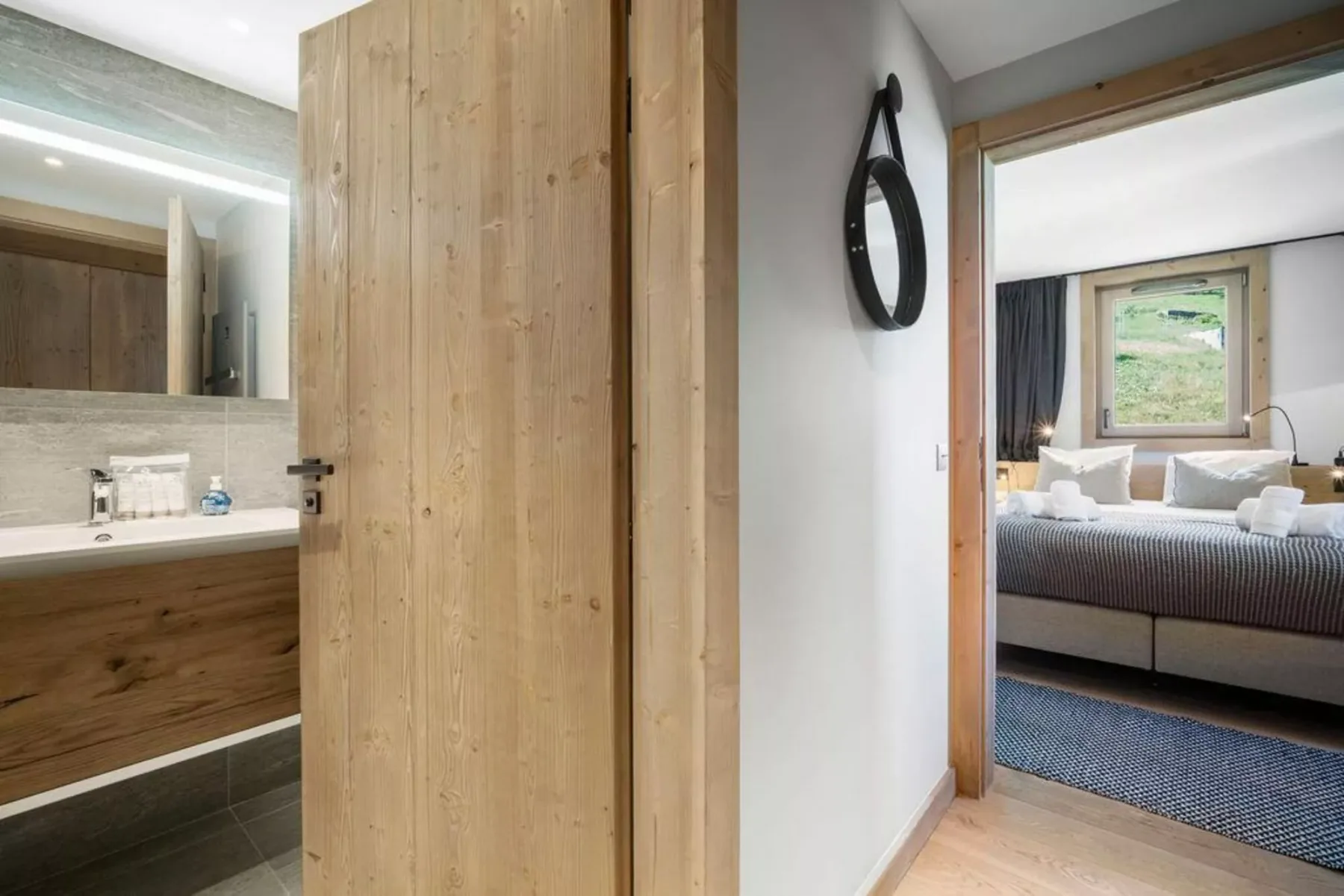 A bathroom vanity and mirror open onto a bedroom with a bed and window.