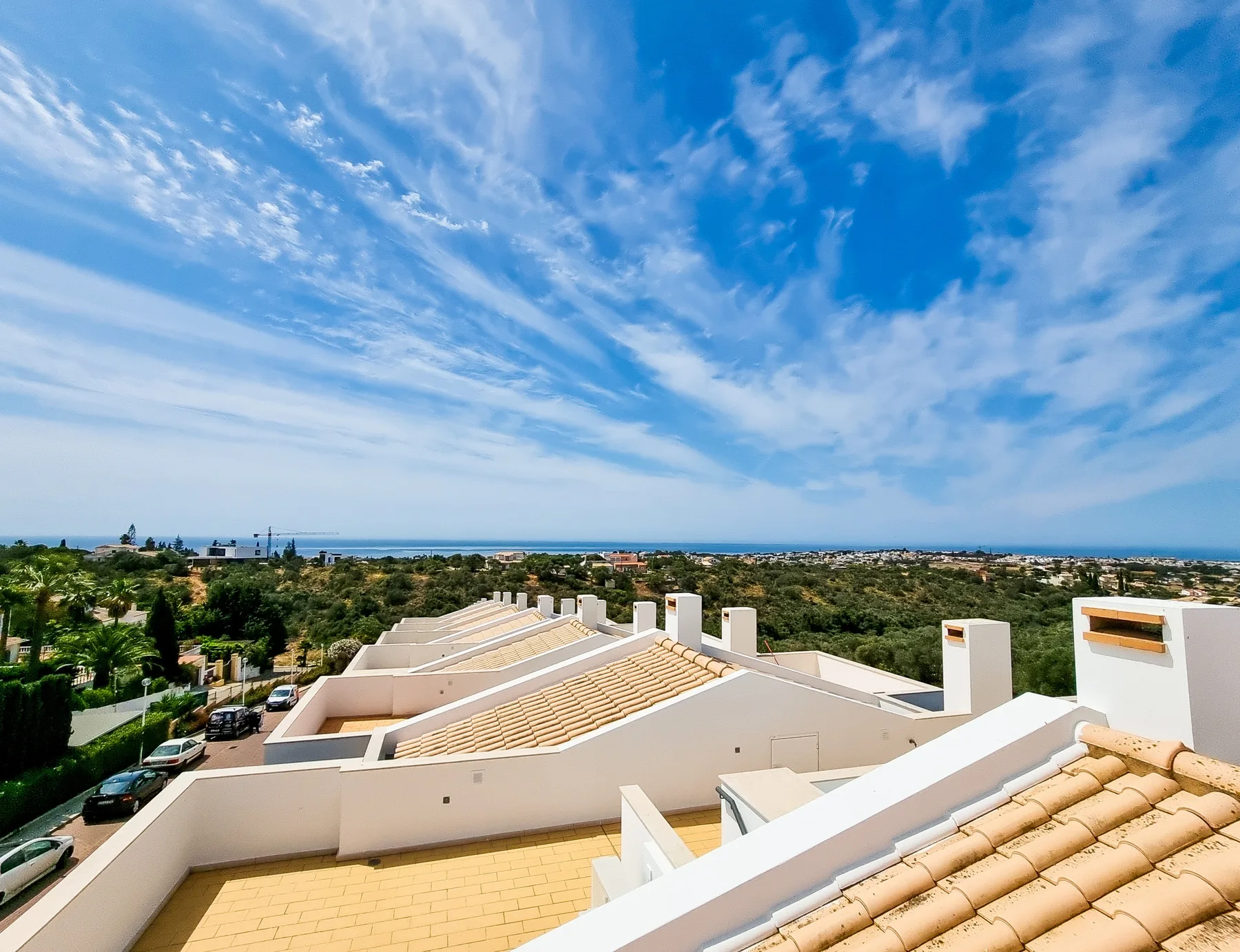 A rooftop terrace overlooks tiled rooftops, greenery, and the ocean beneath a blue sky.