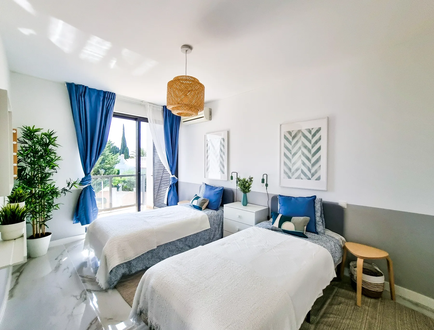 Un dormitorio luminoso con dos camas, cortinas azules y cuadros en la pared.