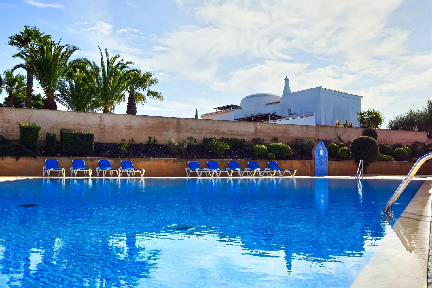 An outdoor swimming pool with blue loungers beside a garden wall and palm trees.