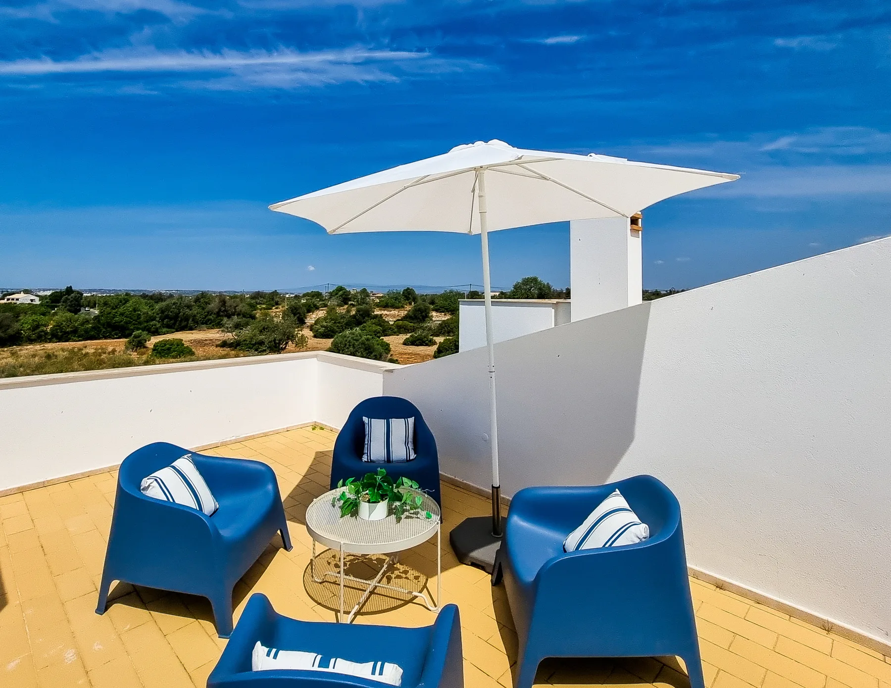 A sunny rooftop terrace has blue seating arranged around a small table beneath an umbrella.