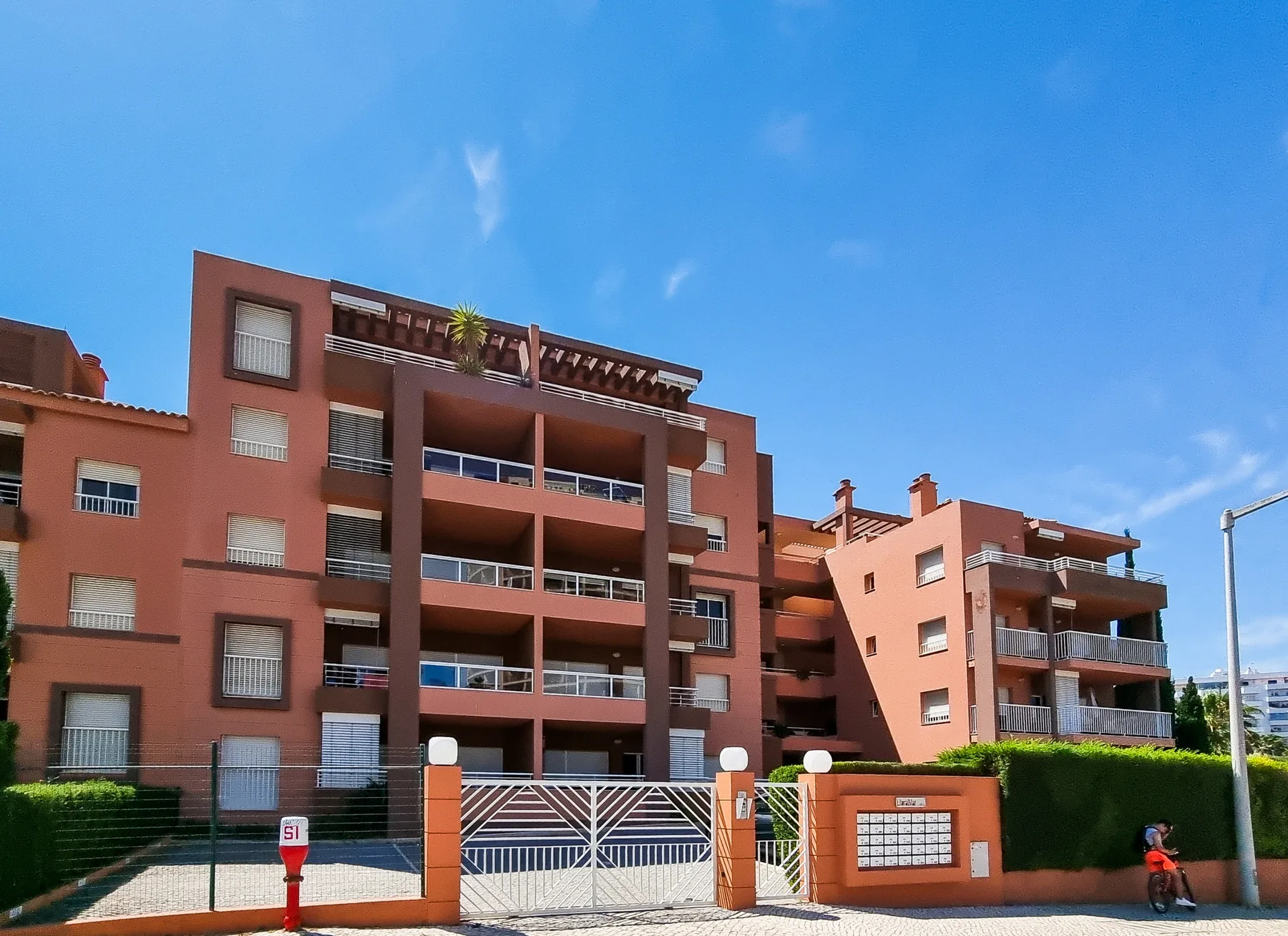 A red apartment building stands behind a gated entrance and trimmed hedges.