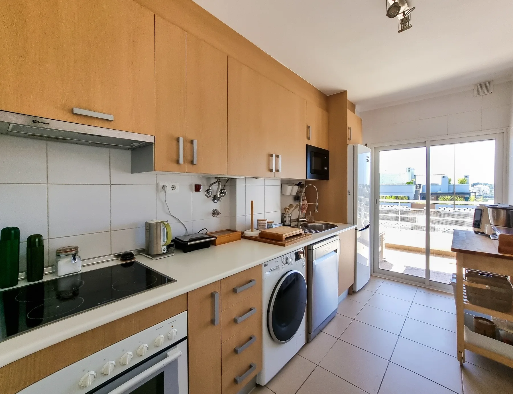 A bright kitchen with wooden cabinets, a cooktop, and large windows opening onto a balcony.
