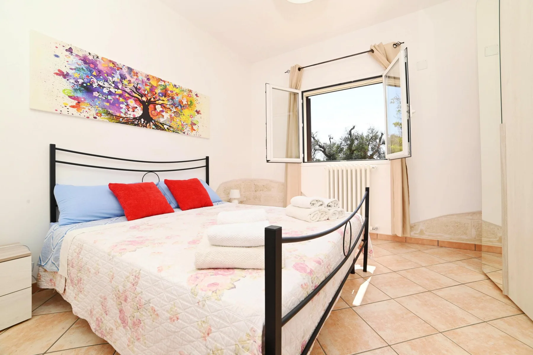 A bright bedroom with a bed beneath colorful wall art and an open window.