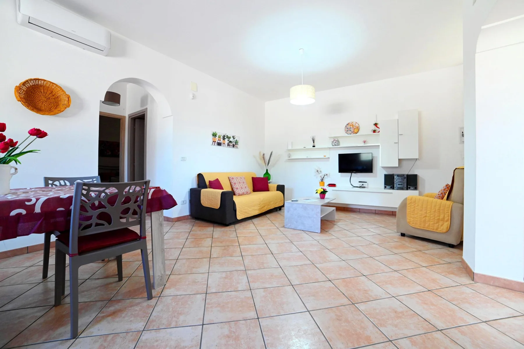 A bright living and dining area with a sofa, television, and tiled floor.