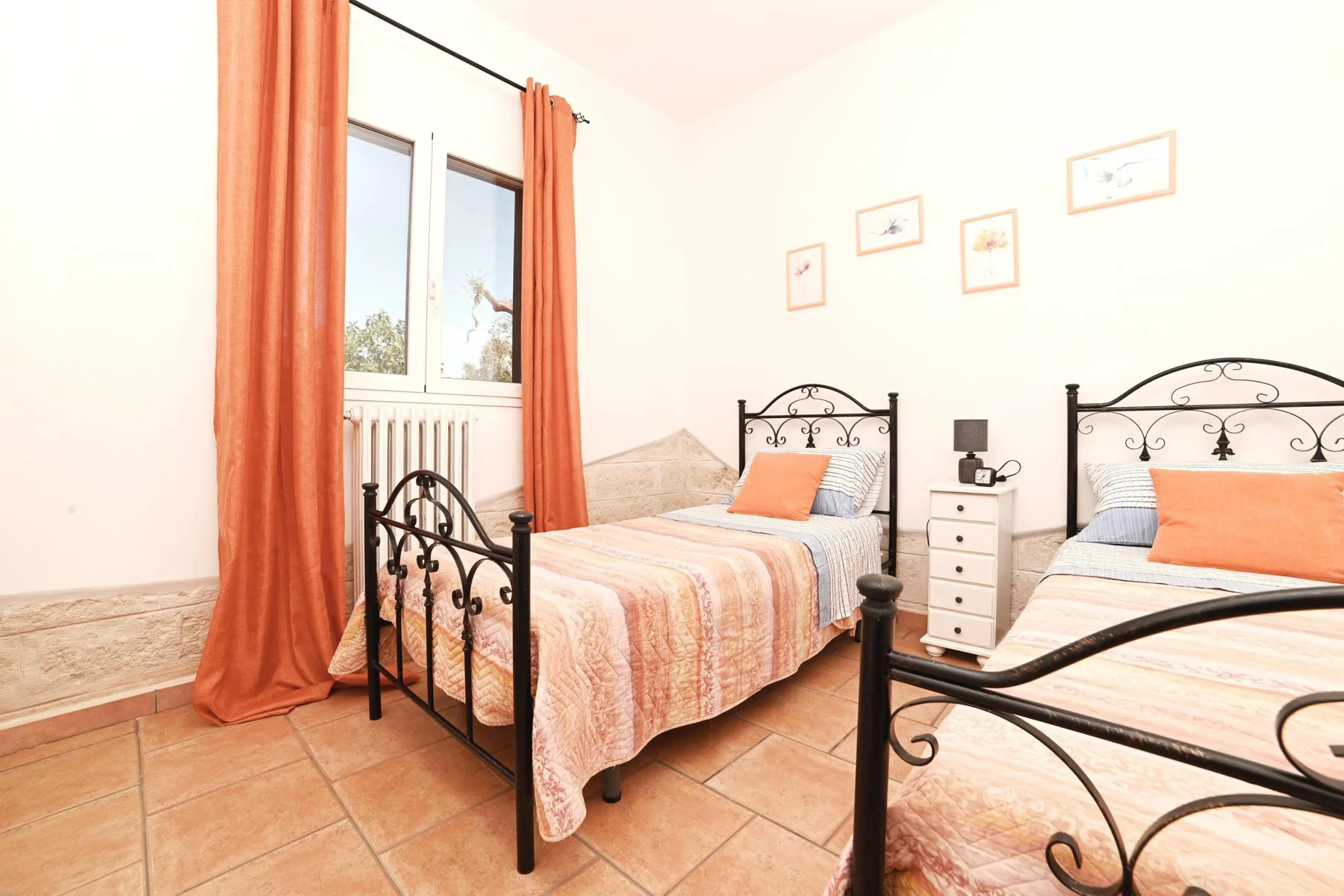 A bedroom with two beds, orange curtains, and framed pictures on the wall.