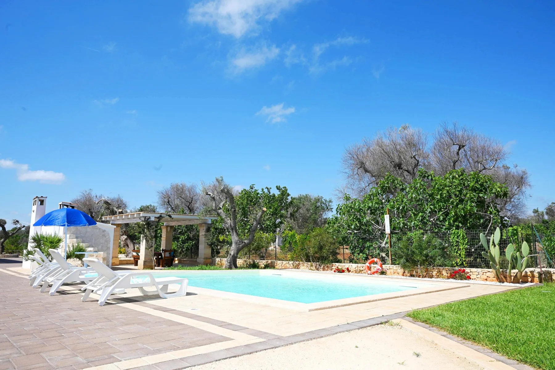An outdoor swimming pool sits beside a paved patio with lounge seating and greenery.