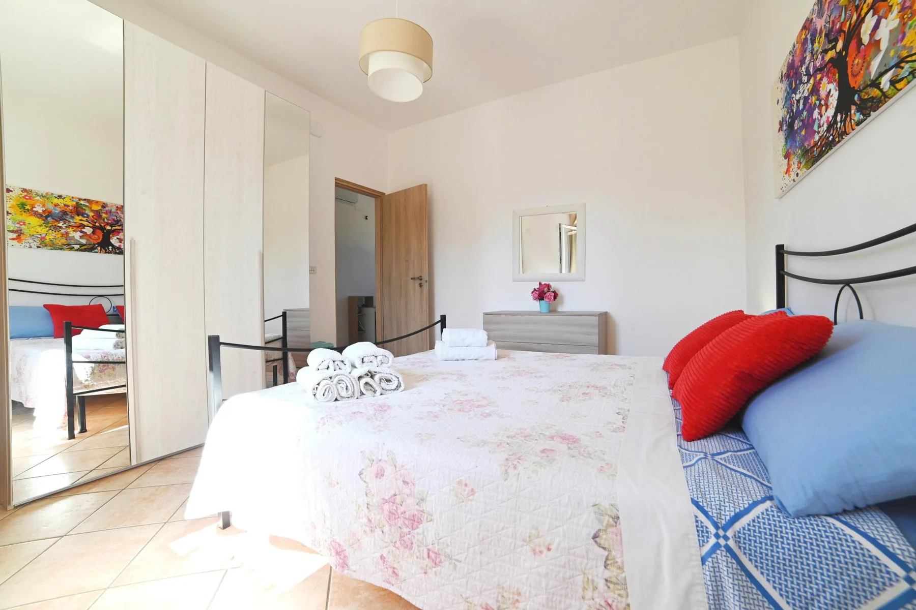 A bright bedroom with a floral bedspread, colorful artwork, and mirrored closet doors.