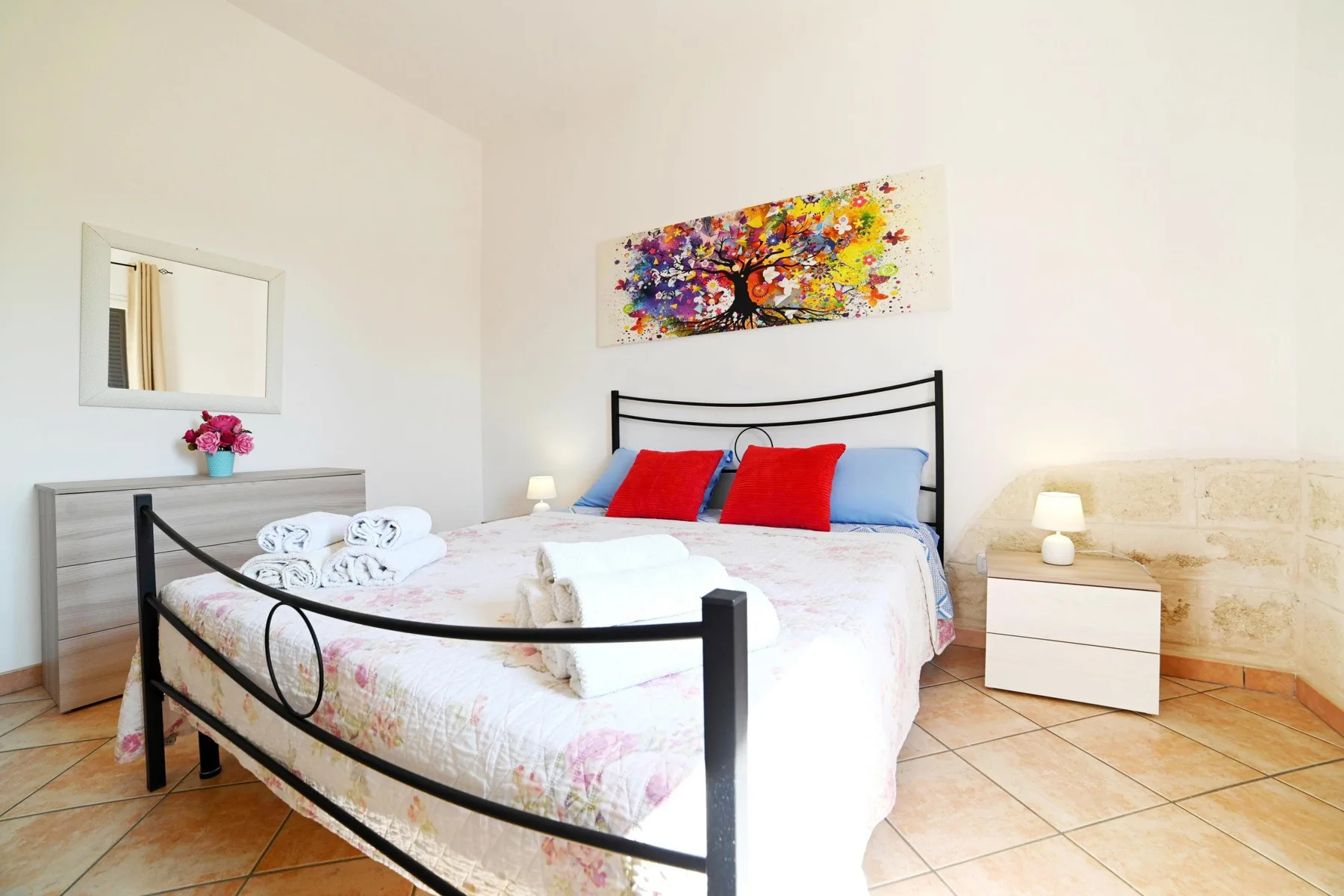 A bright bedroom with a double bed, colorful wall art, and a bedside table.
