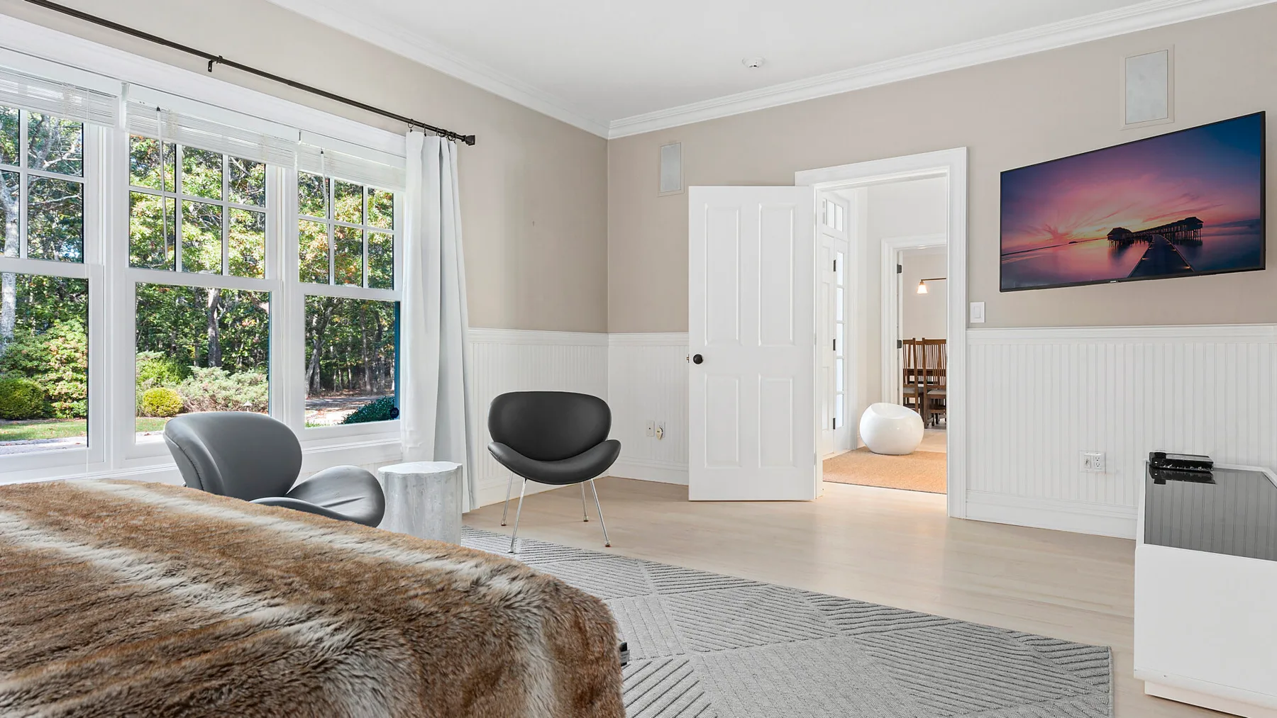 Un dormitorio luminoso con una cama, grandes ventanas y una puerta hacia otra habitación.