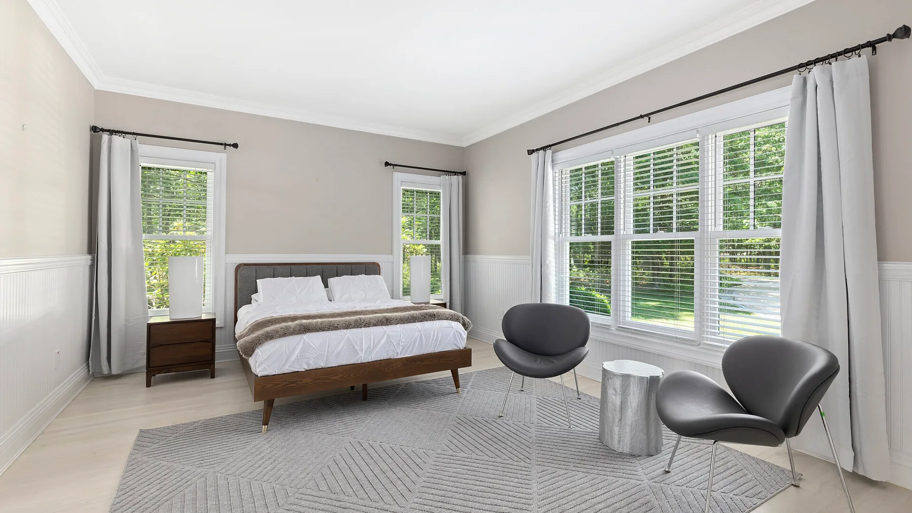 Un dormitorio luminoso con una cama, ventanas amplias y dos sillones.