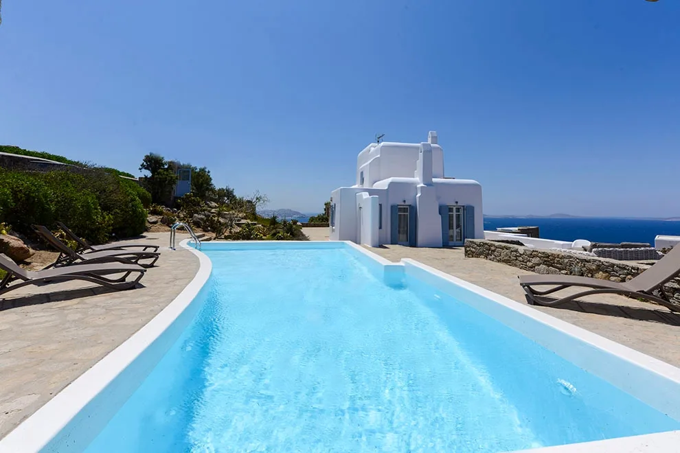 A swimming pool beside a white building, with lounge seating and the sea beyond.
