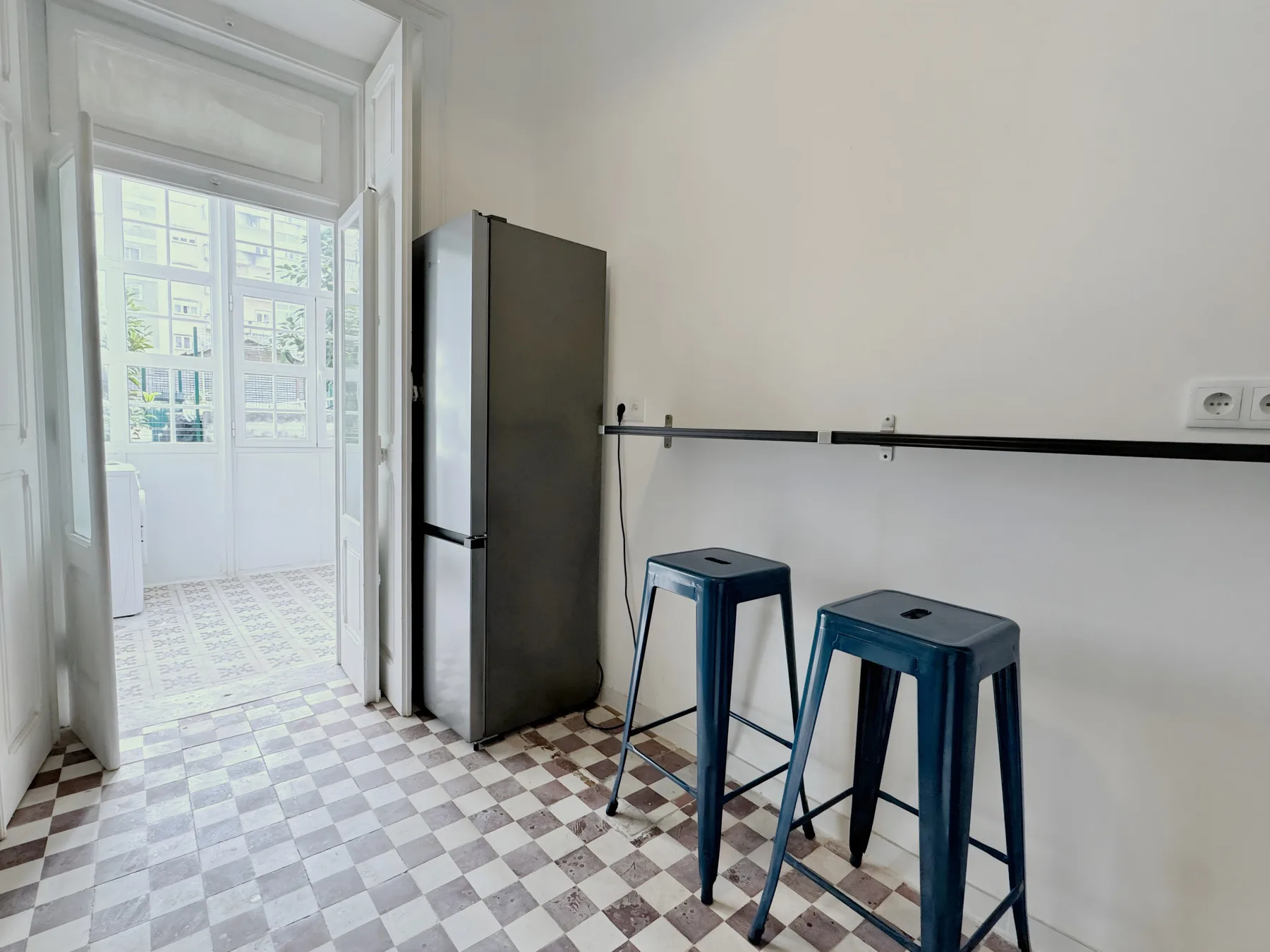 A small kitchen area with a refrigerator and two stools beside a bright adjoining room.