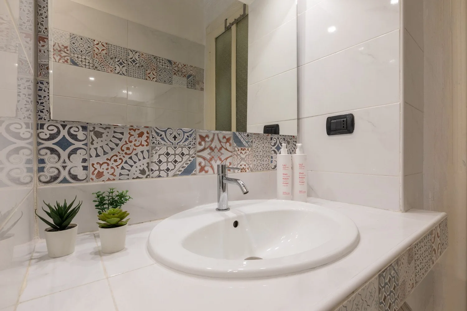 Un lavabo de baño con una pila ovalada, azulejos decorativos y dos pequeñas plantas en macetas.
