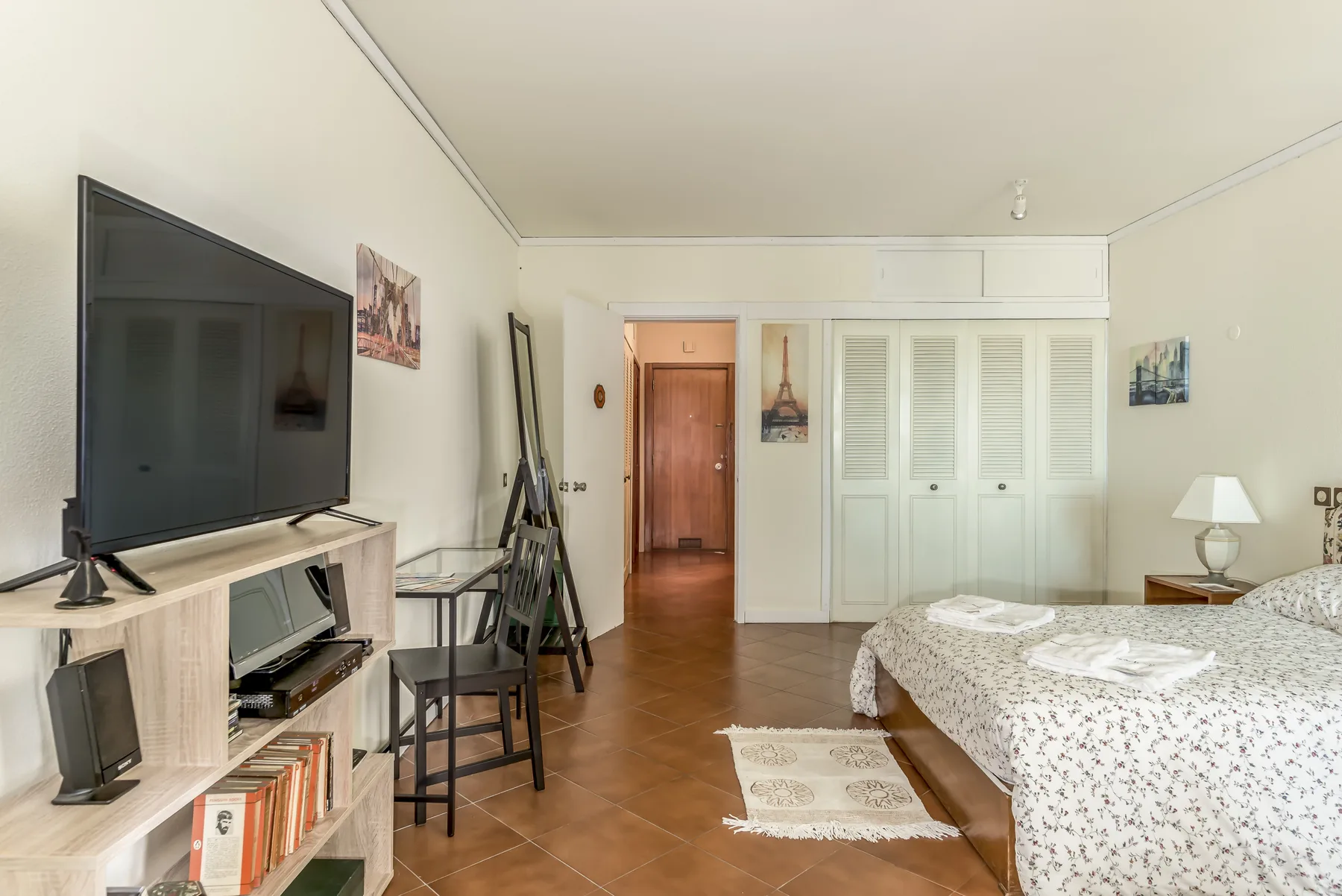 Un dormitorio con televisor, escritorio y cama junto a puertas blancas de armario.