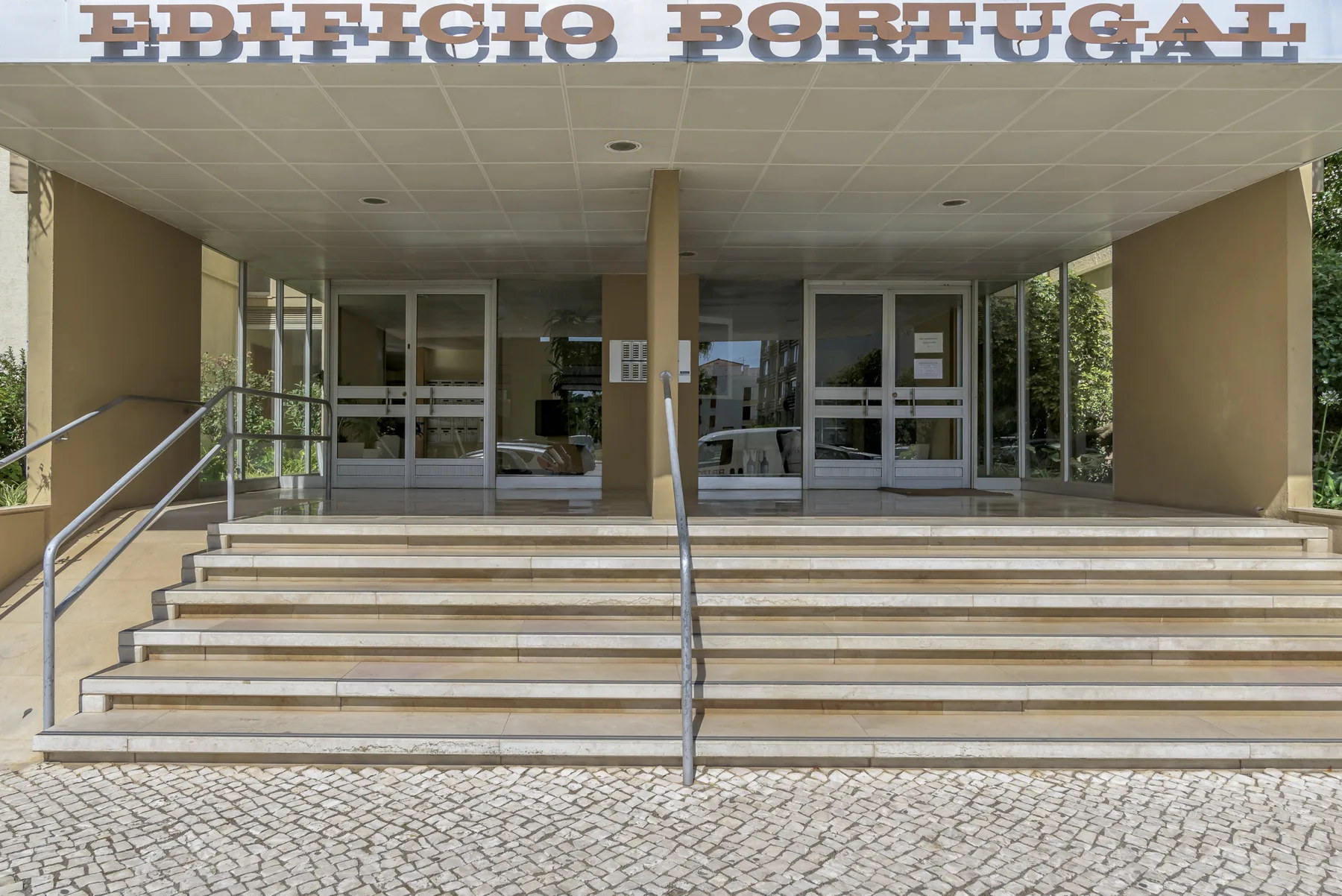 La entrada de un edificio tiene escalones amplios, pasamanos metálicos y puertas de vidrio.