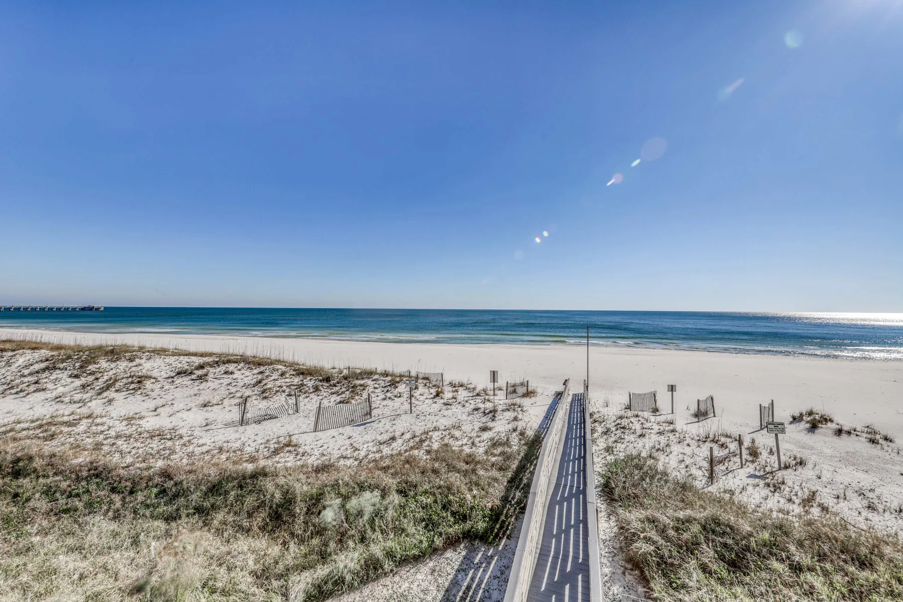 A boardwalk crosses sandy dunes toward the ocean under a clear sky.