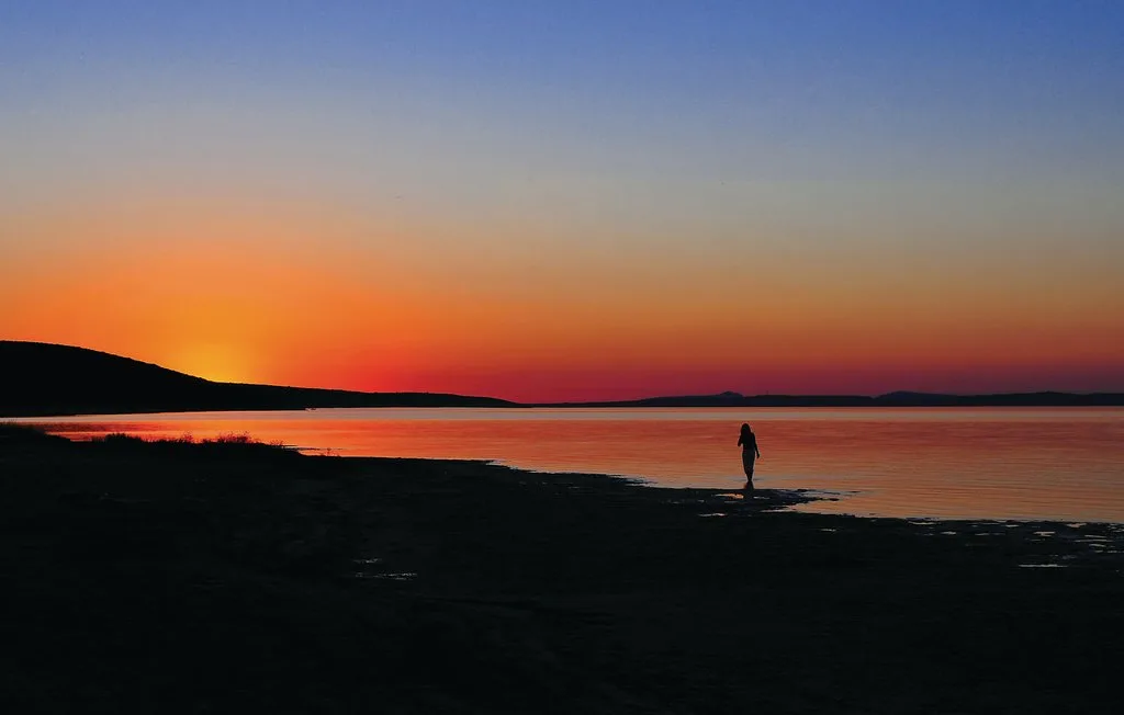 Una persona está junto al agua al atardecer, con una orilla oscura en primer plano.