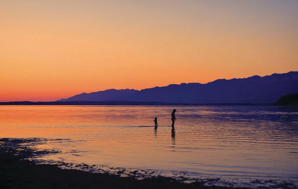 Atardecer sobre aguas tranquilas, con dos figuras de pie cerca de la orilla.