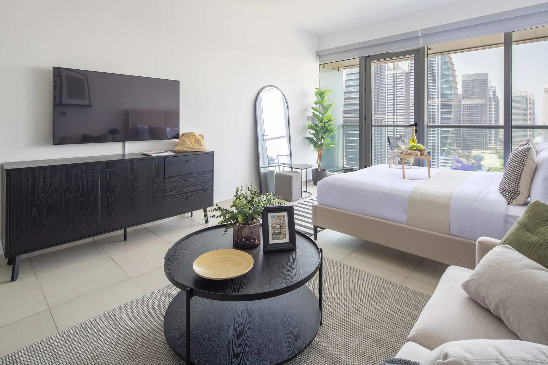 Un dormitorio luminoso con cama, asientos y grandes ventanas con vistas a edificios de la ciudad.
