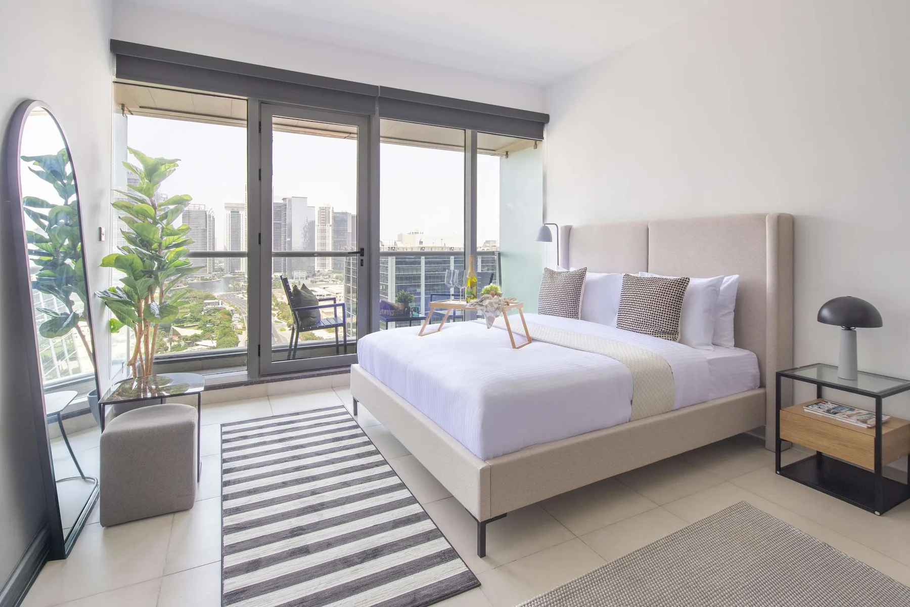 A bright bedroom with a bed, striped rug, and large windows overlooking city buildings.