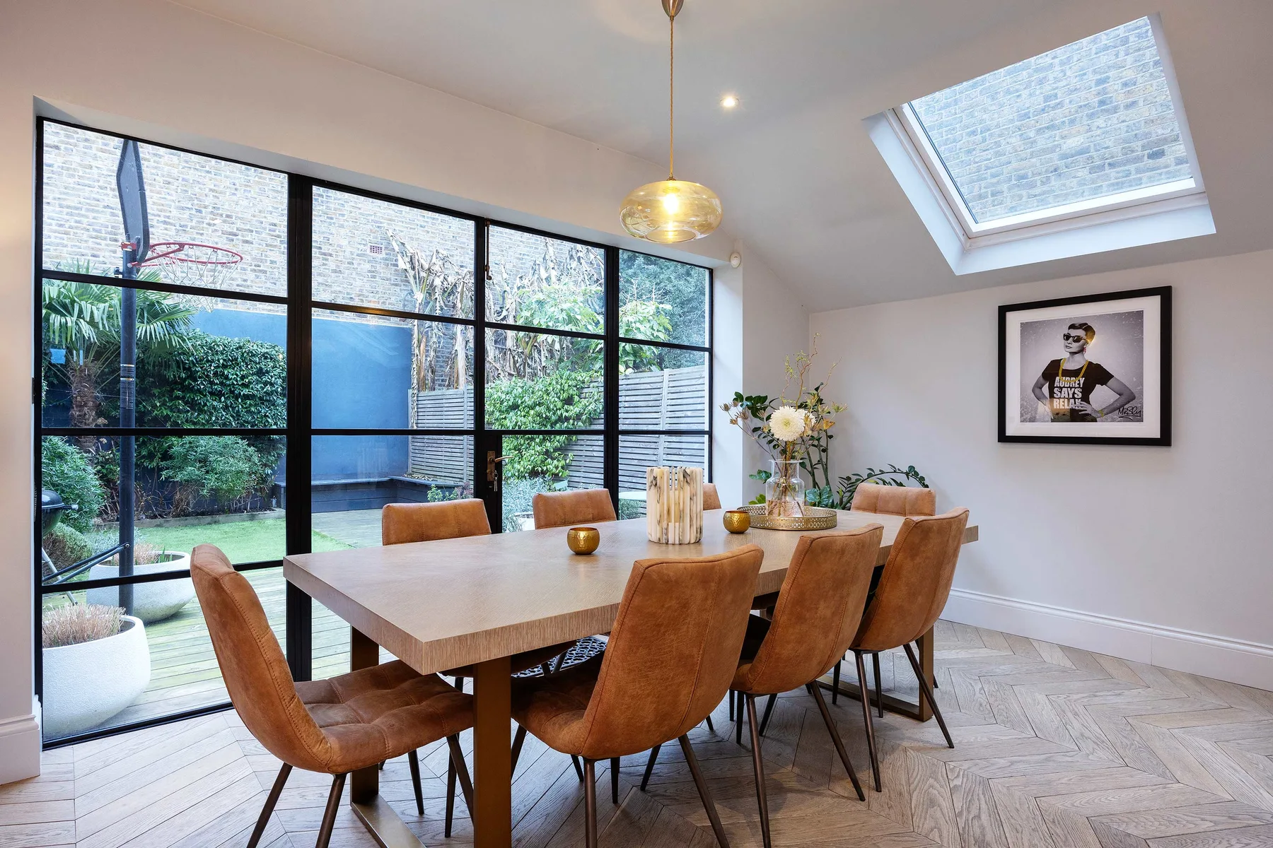 A dining area with a long wooden table beside large windows overlooking greenery.