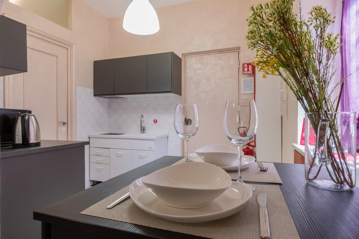 Una mesa de comedor está puesta junto a una cocina compacta, con flores en un jarrón de vidrio.