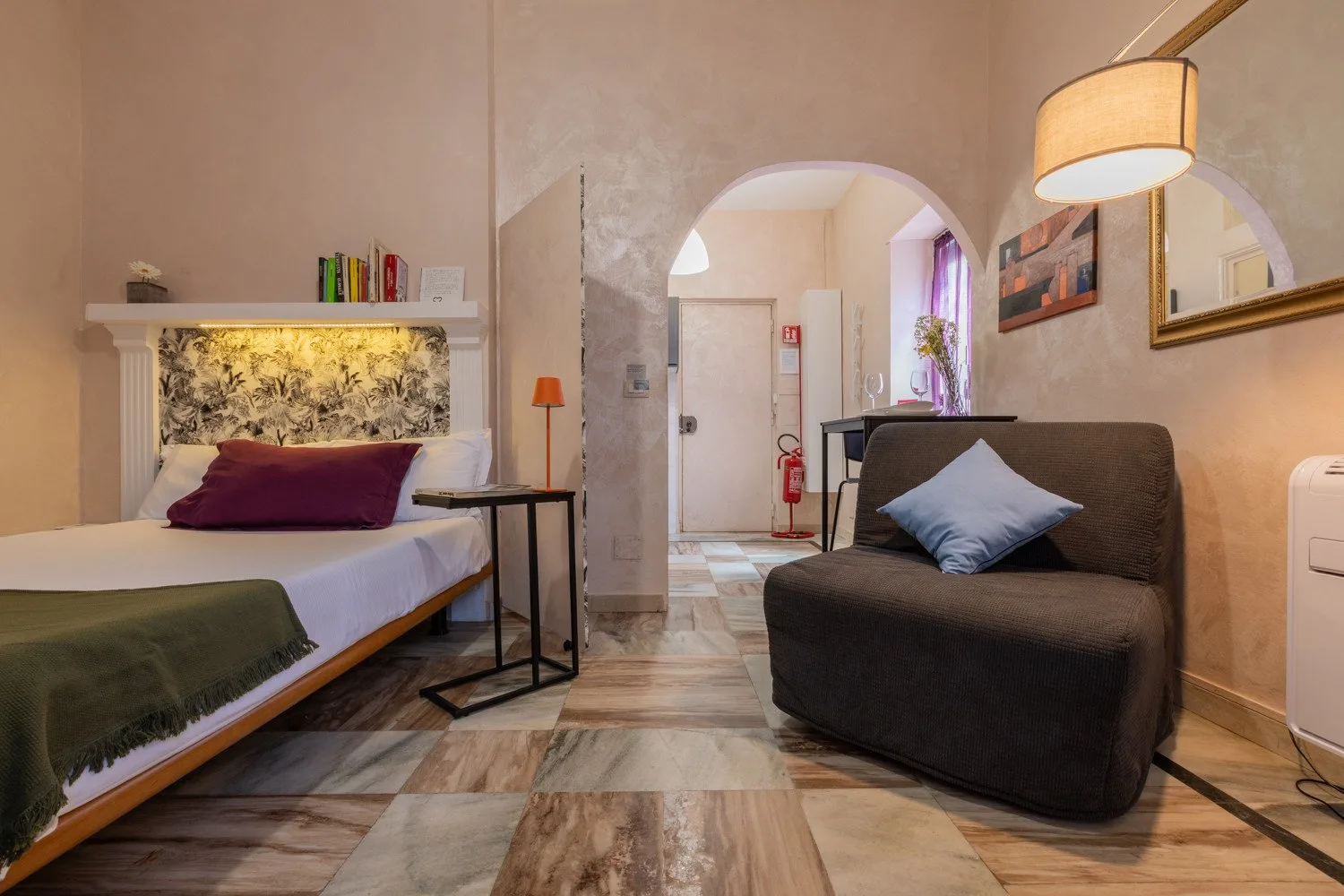 Dormitorio y zona de estar con una cama, asientos oscuros y una puerta arqueada.