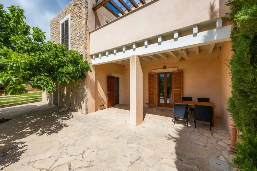 A shaded stone patio sits beside a stucco building, with outdoor seating near the doors.