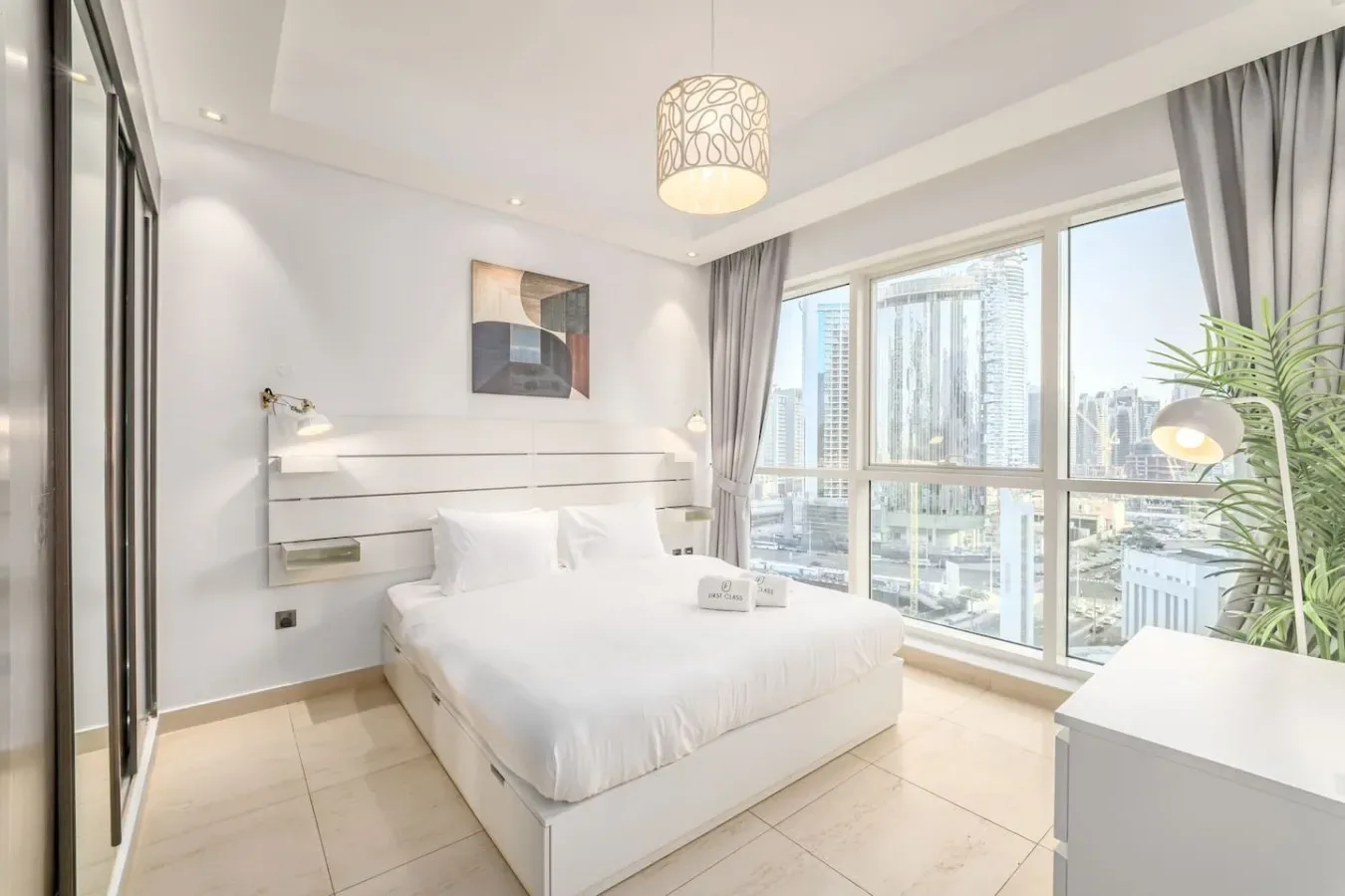 Un dormitorio luminoso con una cama blanca junto a grandes ventanales con vistas a edificios.
