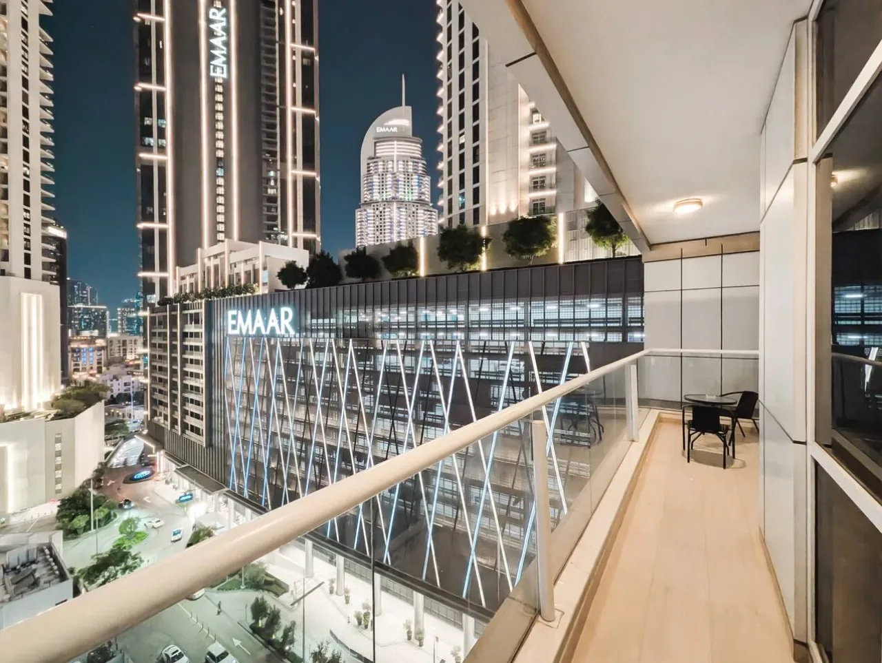 A balcony overlooks illuminated high-rise buildings at night, with a small seating area.