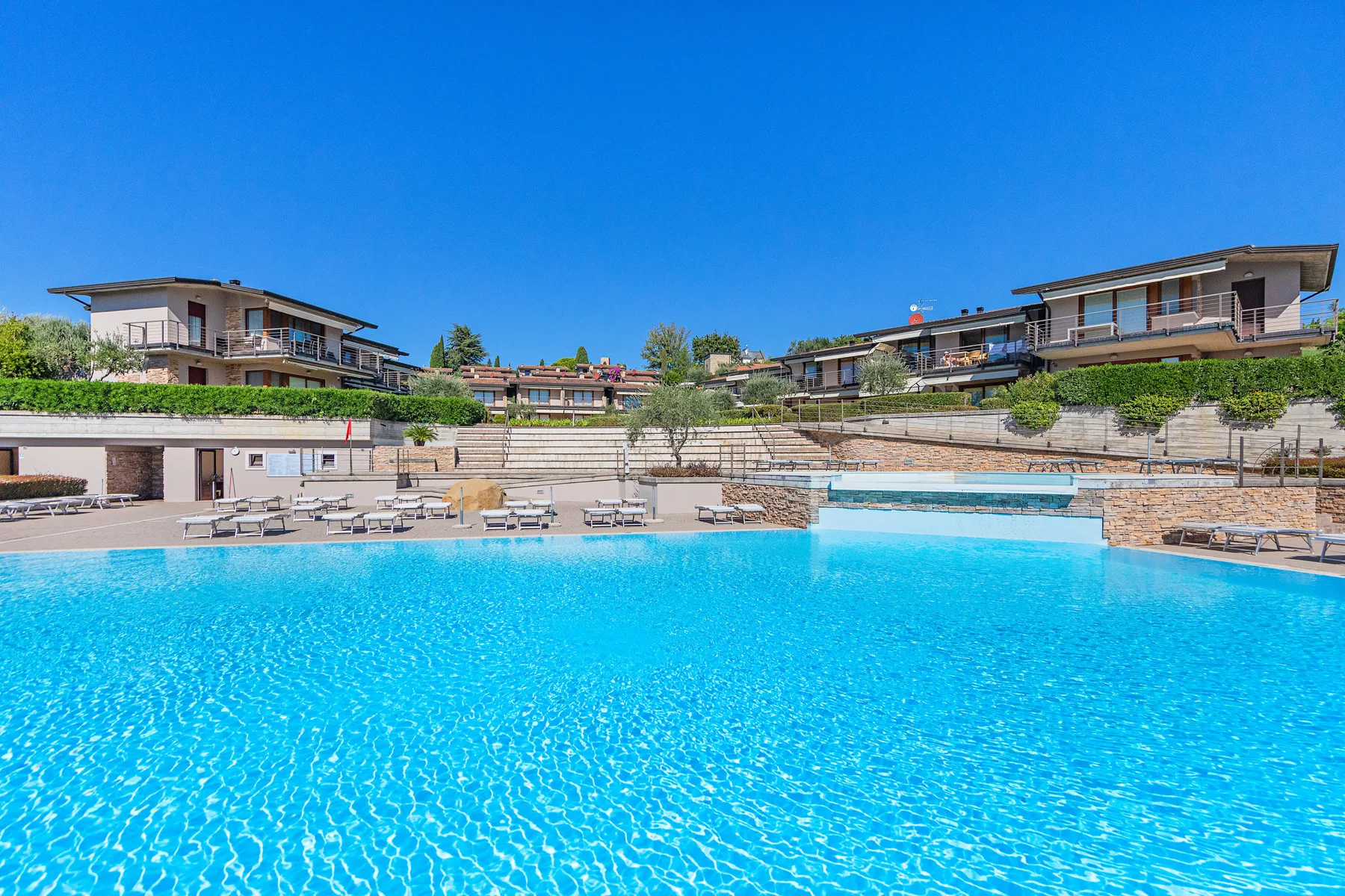 A large outdoor swimming pool sits below terraced buildings under a clear blue sky.