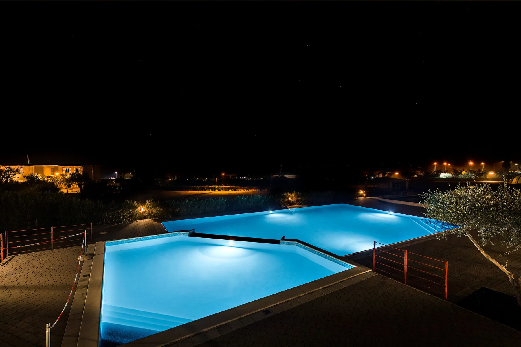 A swimming pool glows blue at night, with lights along the surrounding landscape.