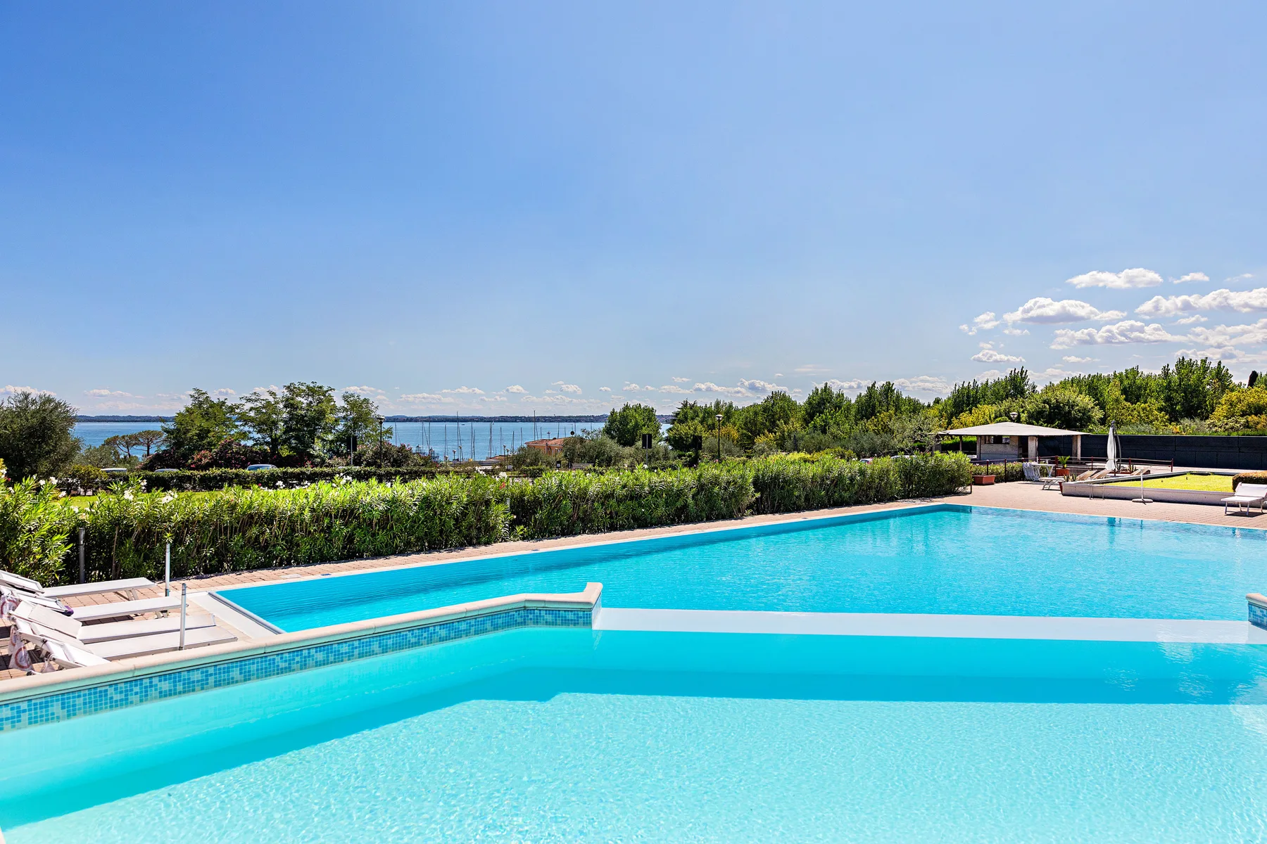 An outdoor swimming pool sits beside landscaped greenery, with water visible beyond.
