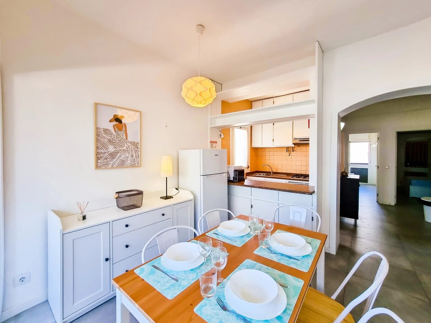 Comedor con mesa de madera y asientos blancos junto a una cocina compacta.