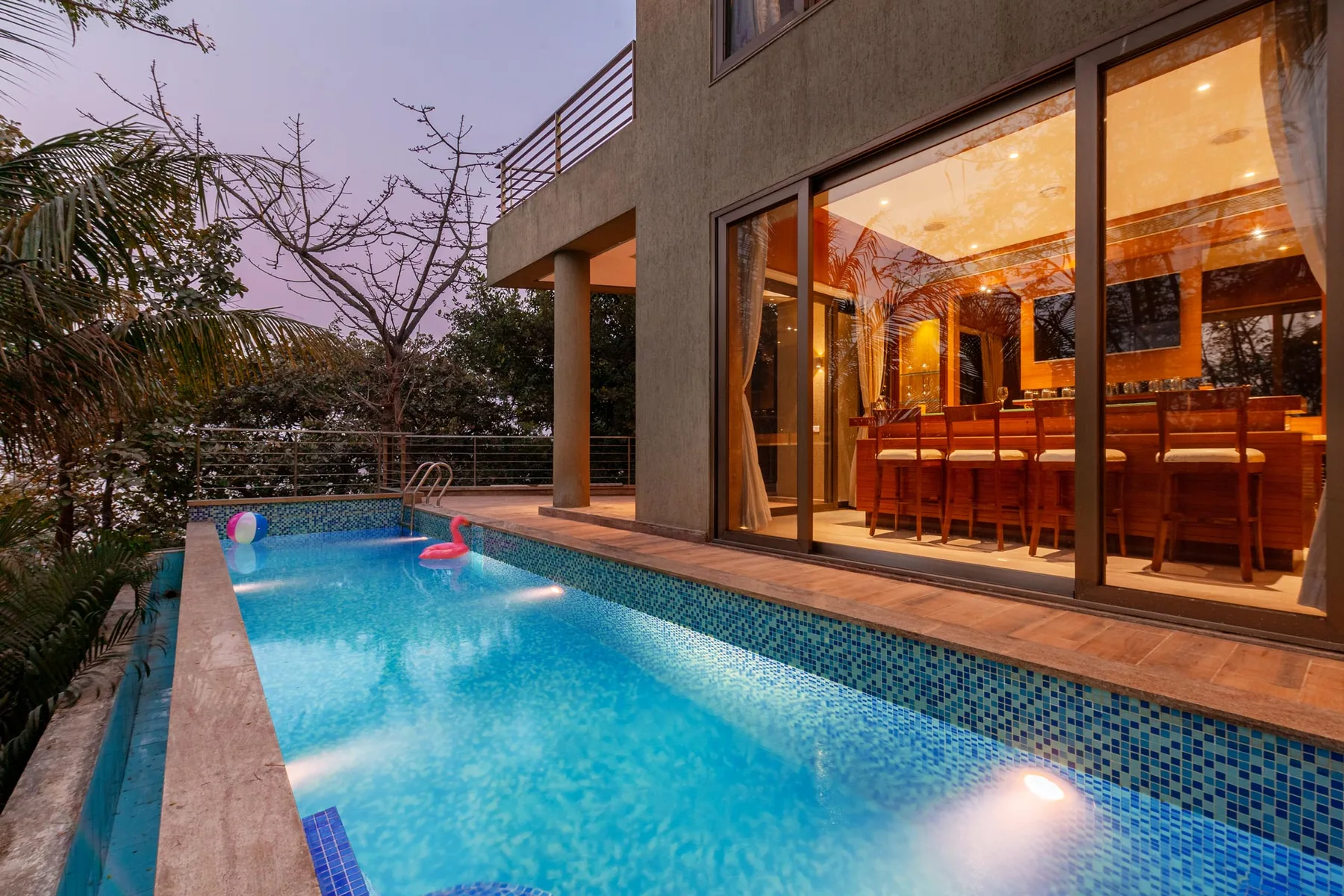 Una piscina junto a una casa moderna, con un comedor iluminado visible a través de puertas de vidrio.