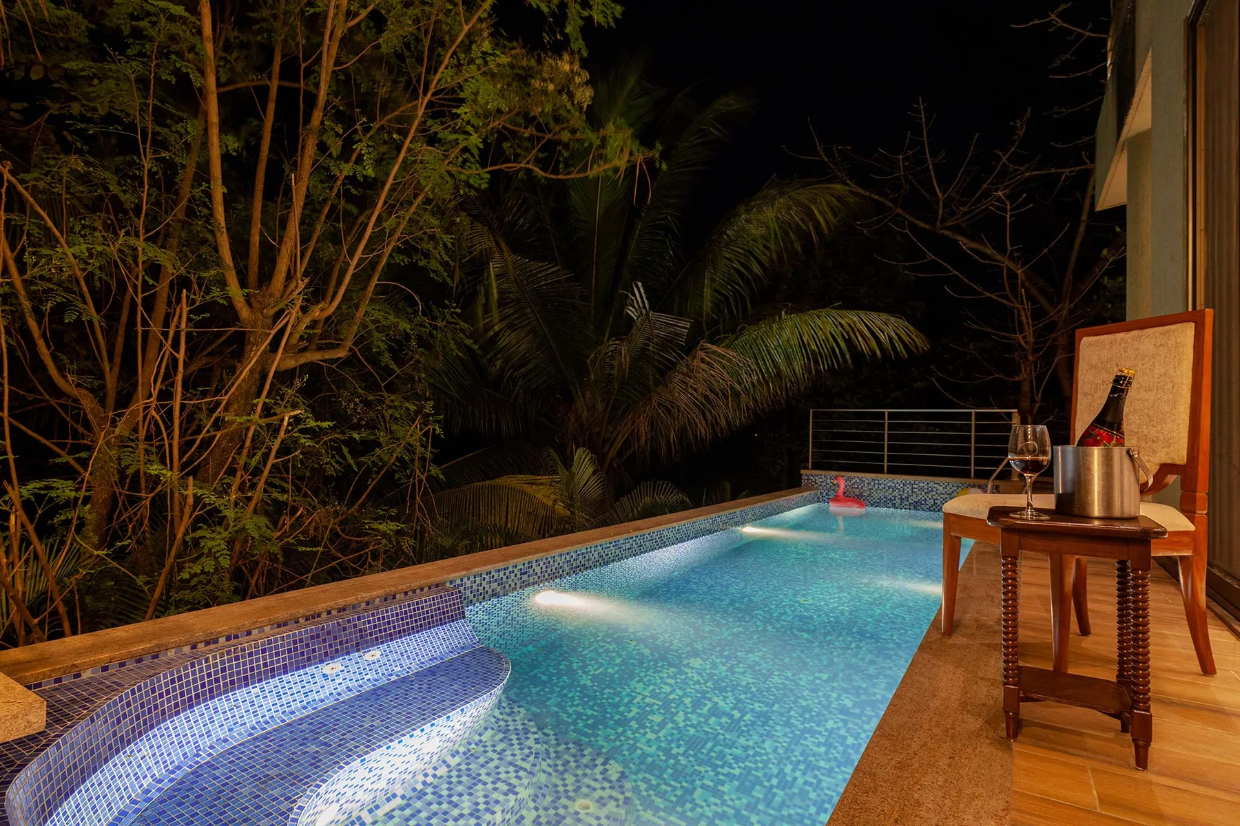 Una piscina de noche está rodeada de árboles tropicales y una terraza de madera.
