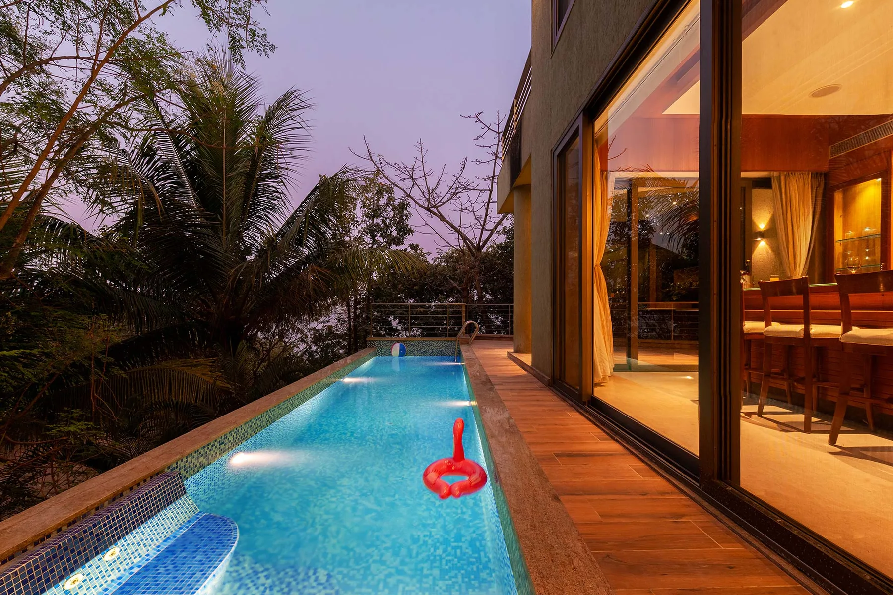 Una piscina junto a una terraza de madera y puertas de vidrio iluminadas.