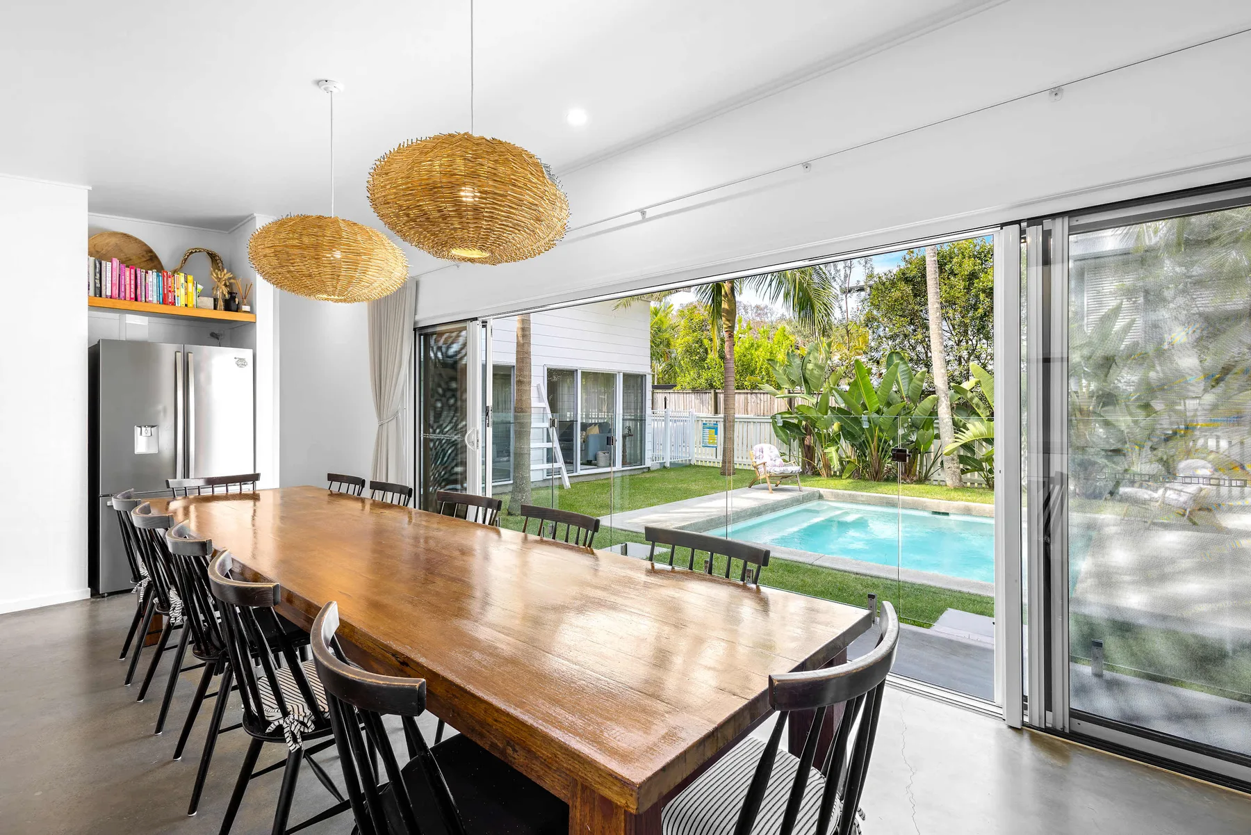 Comedor con una mesa larga de madera junto a puertas de vidrio abiertas con vista a una piscina.
