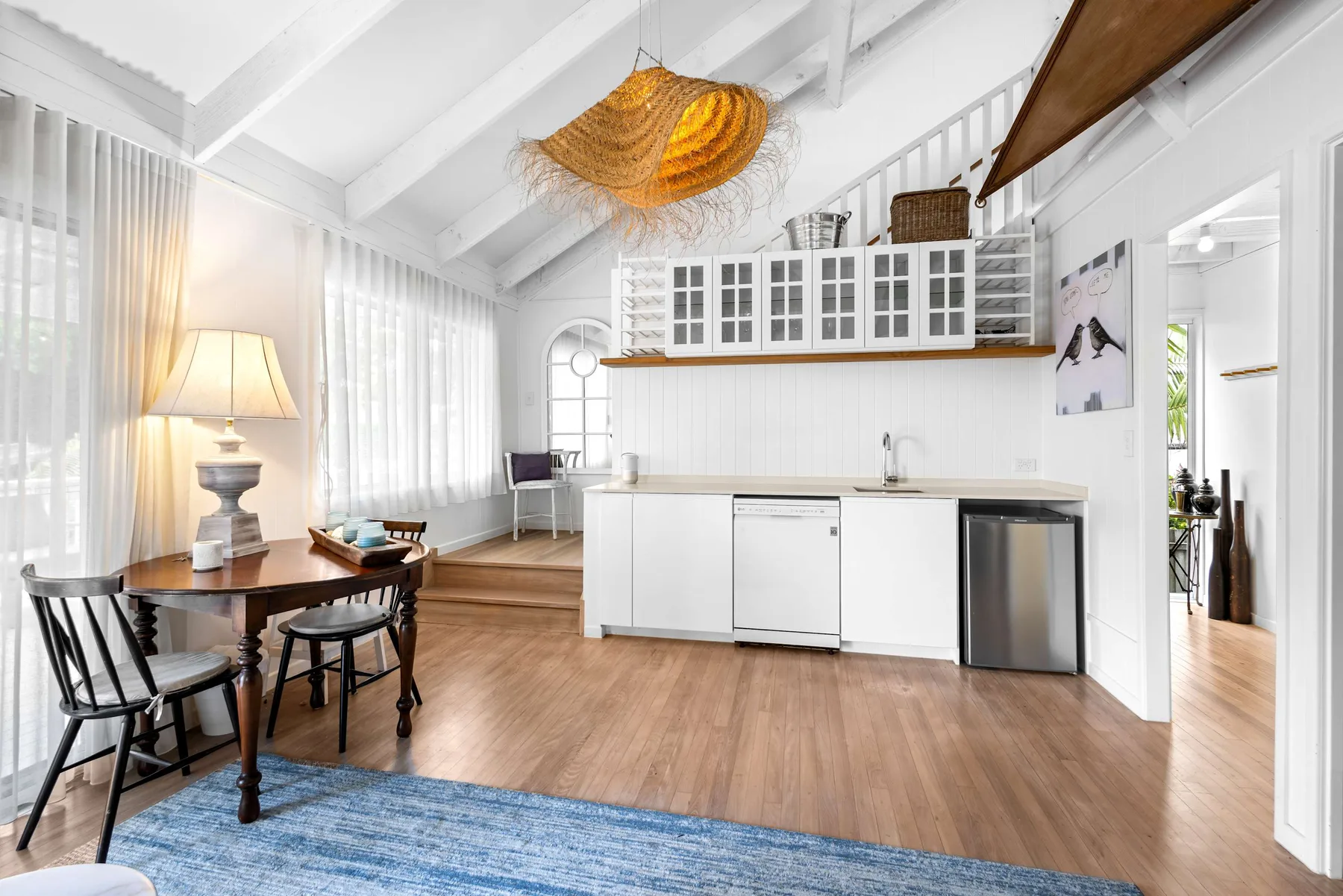 Una luminosa zona de comedor y cocina con una mesa de madera y armarios blancos.
