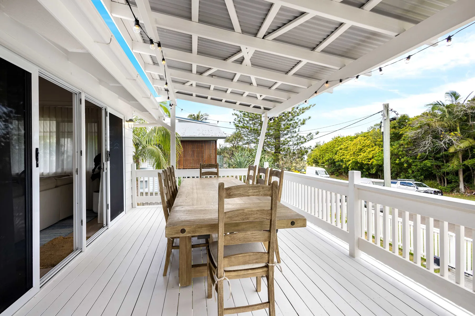 Una terraza exterior cubierta tiene una mesa de comedor de madera y sillas junto a una barandilla blanca.