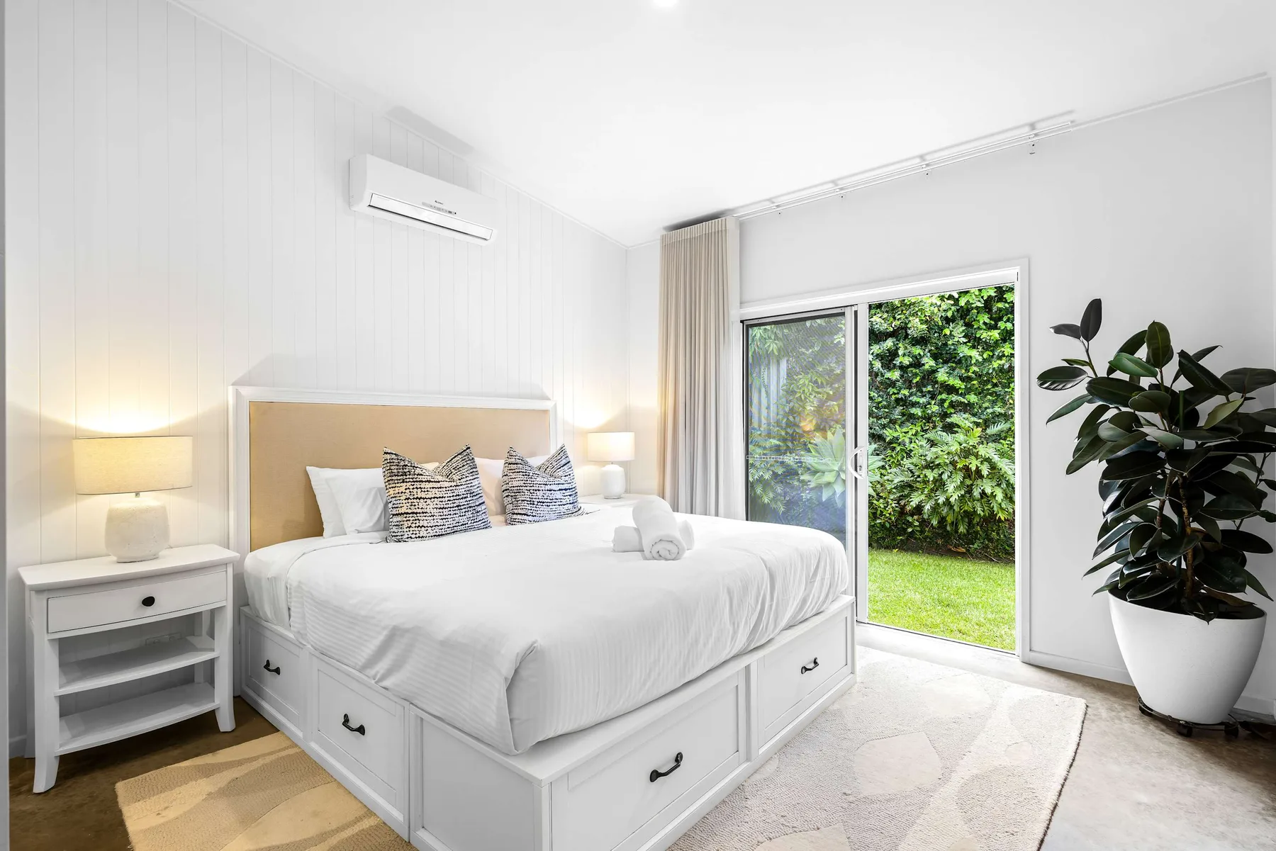 Un dormitorio luminoso con una cama blanca junto a una puerta de vidrio con vistas a la vegetación.